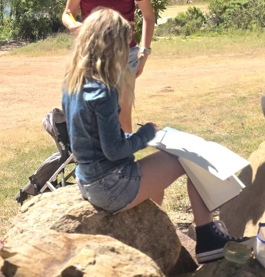 A student perched on a boulder outdoors, sketching in her Field Journal.