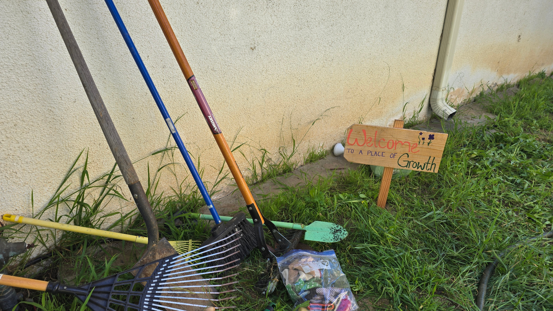 Garden tools leaning against a wall beside a hand-painted 'Welcome to a Place of Growth' sign.