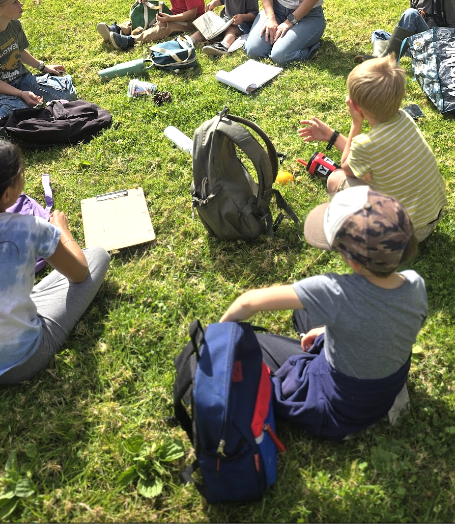 Students seated in a circle on the grass with journals and backpacks during an outdoor lesson.