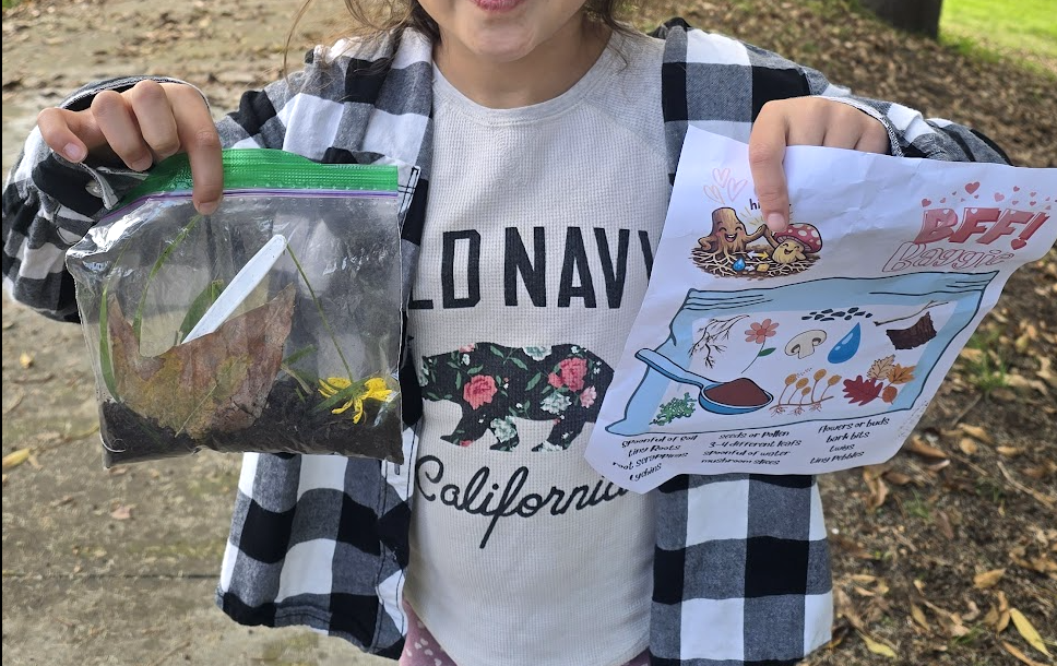 A young student proudly displaying a bag of foraged plants next to a nature scavenger hunt sheet.