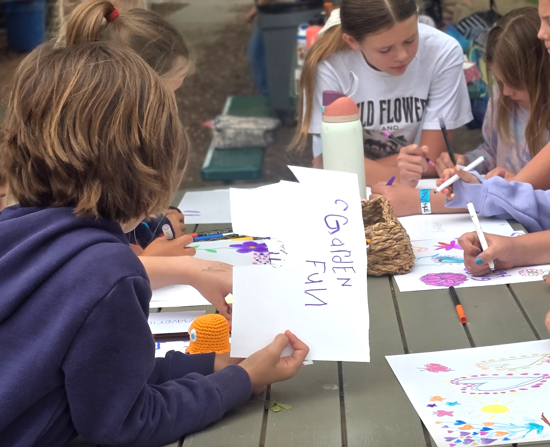 Students drawing colorful flower sketches at an outdoor table, one holding a 'Garden Fun' sign.